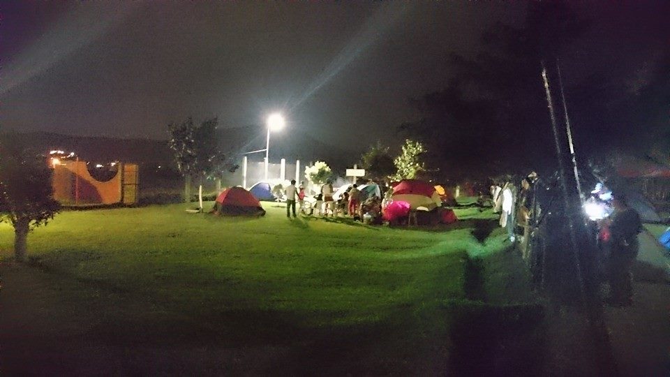 Fotografiá de la zona de campamento, la fotografía se toóo durante la noche, se ve una suelo cubierto de un verde pasto. Al fondo hay casas de campañas y personas conviviendo.