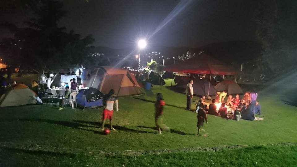 Fotografiá de la zona de campamento, la fotografía se tomó durante la noche, se ven personas jugando con una pelota, mas al fondo hay personas rodeando una fogata, y mas a la izquierda hay casas de campaña y un par de arboles.