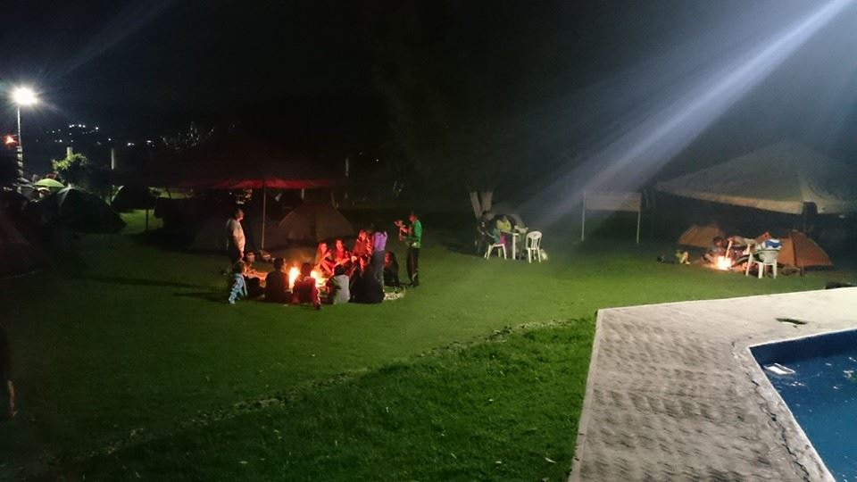 Fotografiá de la zona de campamento, la fotografía se tomó durante la noche, se ven personas conviviendo al rededor de una fogata, mas al fondo, se ven a mas personas con otra fogata y detrás hay casas de campañas y un gran árbol.