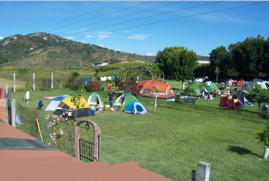Fotografiá de la zona de campamento, la fotografía se tomó durante el día, la fotografía se tomó desde el techo de una cabaña. Se ve la zona de acampar, cubierta por un verde pasto, hay muchas casas de campaña y algunos arboles al fondo.