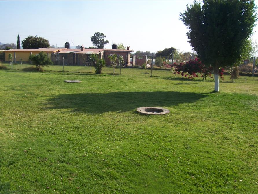 Fotografiá de la zona de campamento, la fotografía se tomó durante el día. Se ve un verde pasto que cubre el suelo, un par de huecos hechos con semento separados entre si, son usados para encender la fogata dentro de ellos y no dañar el suelo. Al fondo, se un árbol y las cabañas del balneario.