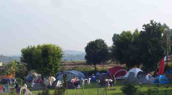 Fotografiá de la zona de campamento, la fotografía se tomó durante el día. Se ve muchas casas de campaña, un verde parto que cubre el suelo, algunos arboles y personas conviviendo.